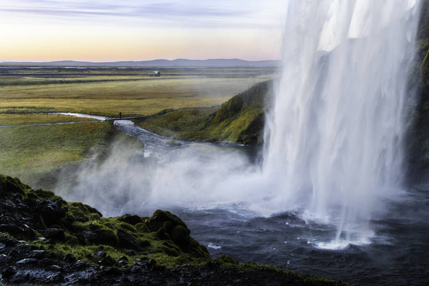 A waterfall is flowing into a river in a grassy field. The water is misty and the sky is a beautiful orange and pink color. The Golden Circle, Iceland. - Photo, Image