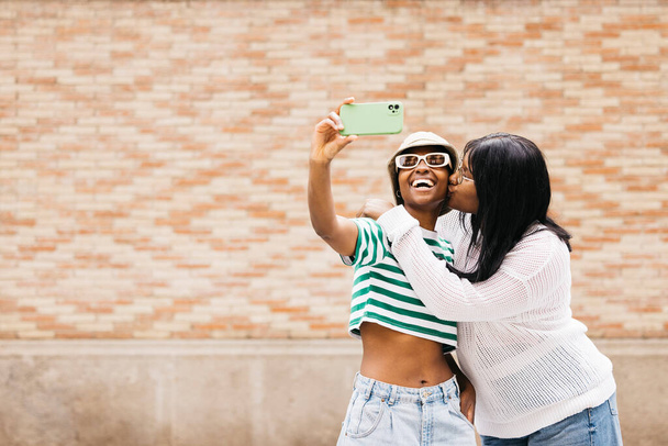 Портрет двух молодых веселых черных женщин, целующихся и делающих селфи на открытом воздухе. - Фото, изображение