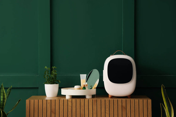 Réfrigérateur cosmétique avec produits, miroir et plante d'intérieur en pot sur commode en bois près du mur vert - Photo, image