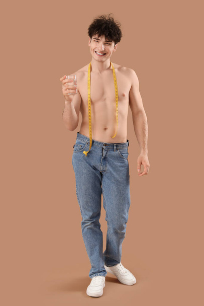 Knappe jonge sportieve man met meetlint en glas water op beige achtergrond. Gewichtsverlies concept - Foto, afbeelding
