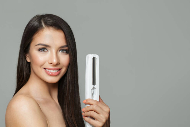 Gorgeous young healthy brunette woman holding hair iron and straightening her long smooth shiny hair on white background. Haircare, hairstyle, hairdressing and hair styling concept - Photo, Image