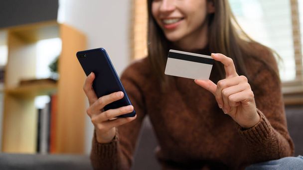 Ευτυχισμένος νεαρός καυκάσιος γυναίκα κρατώντας πιστωτική κάρτα και ψώνια σε απευθείας σύνδεση στο smartphone. - Φωτογραφία, εικόνα