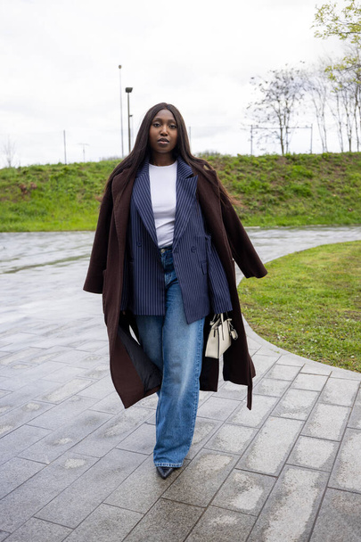 Esta atractiva foto muestra a una joven mujer negra dando un paseo enérgico en un entorno de parque urbano. Shes estilo en un conjunto de otoño de varias capas, con una llamativa chaqueta de rayas de alfiler debajo de un abierto - Foto, imagen