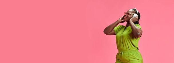 Banner. Joyful woman in vibrant green clothes listening music and dancing against pastel pink studio background with negative space. Concept of human emotions, fashion and beauty, trends. - Photo, Image