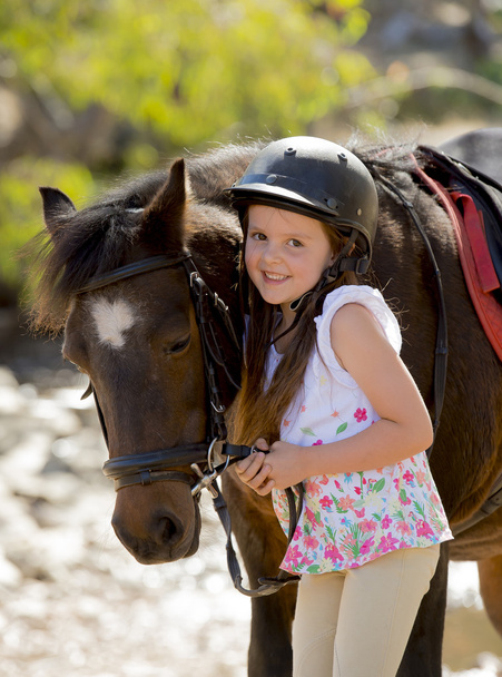 νεαρό κορίτσι 7 ή 8 ετών εκμετάλλευση χαλινάρι μικρό πόνυ αλόγου, χαμογελά ευτυχισμένος φορώντας jockey κράνους ασφαλείας σε καλοκαιρινές διακοπές - Φωτογραφία, εικόνα