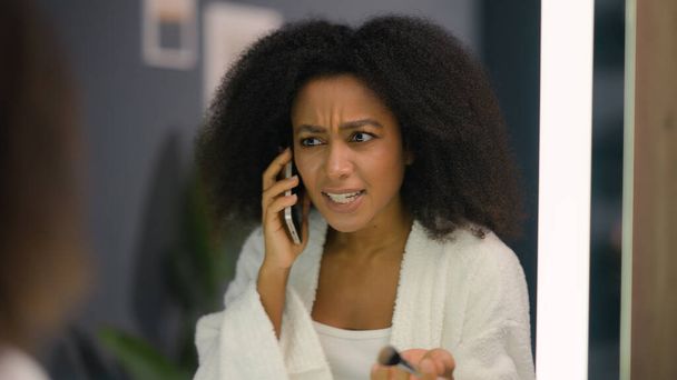 Öfkeli duygusal hüsran stresli Afrikalı Amerikalı kadın rahatsız olmuş etnik kız pudrası organik kozmetik cilt bakımı ev banyosunda aynaya bakarak konuşan telefon çatışması - Fotoğraf, Görsel