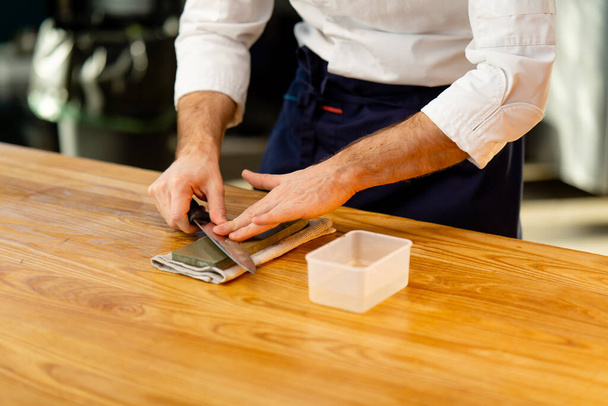 κοντινό πλάνο ενός σεφ με λευκό σακάκι στην κουζίνα στο τραπέζι ακονίζει ένα μαχαίρι στην πράσινη σόμπα - Φωτογραφία, εικόνα