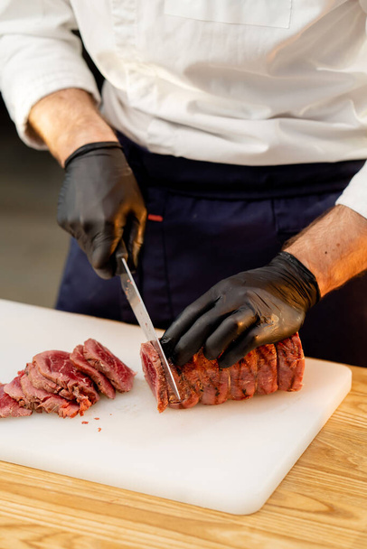 közelkép egy szakács kezét fekete kesztyű vágás egy darab nyers szárított hús egy fórumon - Fotó, kép