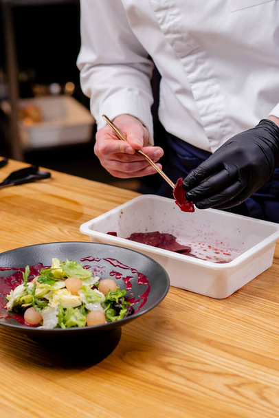 крупным планом черной тарелки, в которой салат повара руками добавляют кусочки свеклы с пинцетом - Фото, изображение