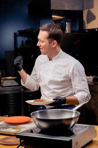 een chef-kok in een professionele keuken staat met een witte plaat van een kant-en-klaar visgerecht verblijdt zich en danst - Foto, afbeelding