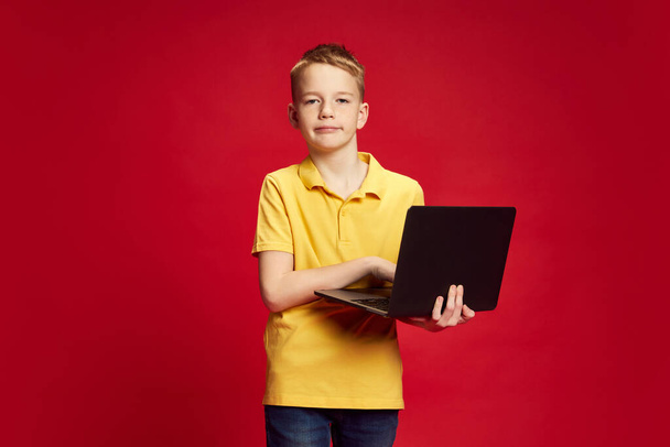 Πολύ διάβασμα για το σπίτι. Εφηβικό αγόρι, ντυμένο casual ντύσιμο με βαρετή έκφραση στην κάμερα σε φόντο κόκκινο στούντιο. Έννοια των ανθρώπινων συναισθημάτων, παιδική ηλικία, εκπαίδευση, μόδα και στυλ. ΠΑΡΑΡΤΗΜΑ II - Φωτογραφία, εικόνα