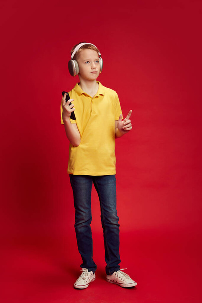 Full-length portrét teenagera, tanec při poslechu hudby ve sluchátkách na pozadí červeného studia. Koncept lidských emocí, dětství, vzdělání, módy a stylu. Reklama - Fotografie, Obrázek