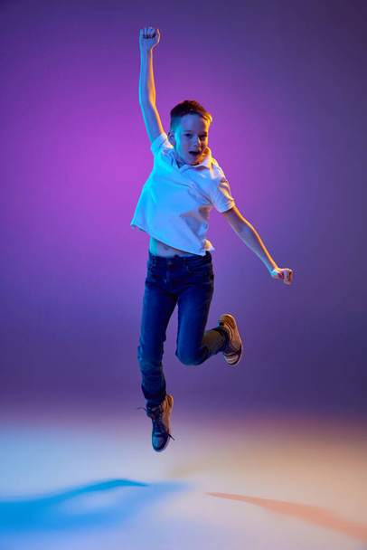 Summer break. Young boy in casual outfit jumping raising hands of joy and fun in neon light against purple gradient background. Concept of human emotions, childhood, education, fashion and style. Ad - Photo, Image