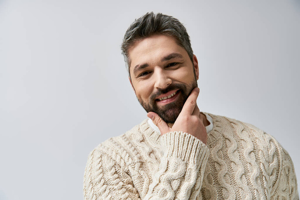 Een boeiende man met een baard die in een witte trui tegen een grijze studio-achtergrond poseert. - Foto, afbeelding