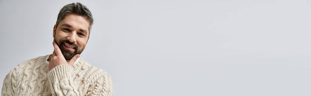 Charismatický muž s vousy zaujme pózu v bílém svetru na neutrálním šedém pozadí ve studiu. - Fotografie, Obrázek