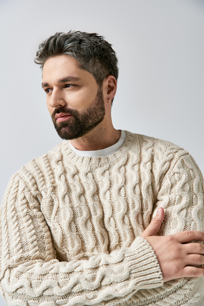 Un homme à la mode avec une barbe frappe une pose dans un pull blanc sur un fond de studio gris. - Photo, image