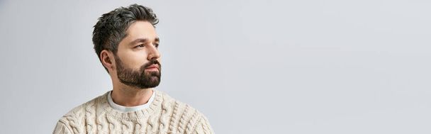 Харизматичный человек с бородой наносит позу в уютном белом свитере на фоне серой студии. - Фото, изображение