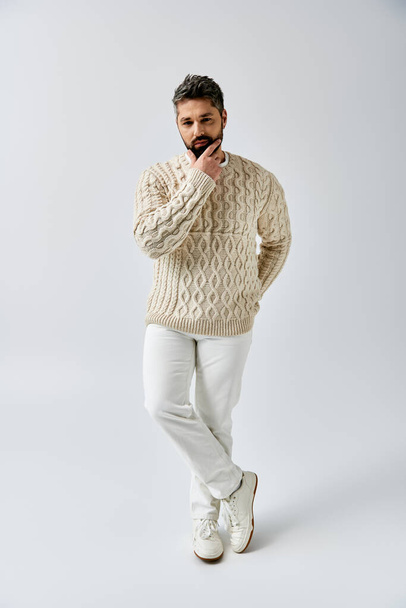 Ένας γενειοφόρος άνδρας ποζάρει με χάρη σε ένα λευκό πουλόβερ σε ένα γκρι φόντο στούντιο. - Φωτογραφία, εικόνα
