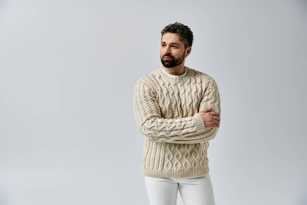Ein stilvoller Mann mit Bart posiert im weißen Pullover vor grauer Studiokulisse. - Foto, Bild
