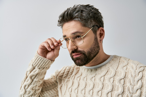 Un homme charismatique avec une barbe pose en pull blanc sur fond gris dans un décor studio. - Photo, image