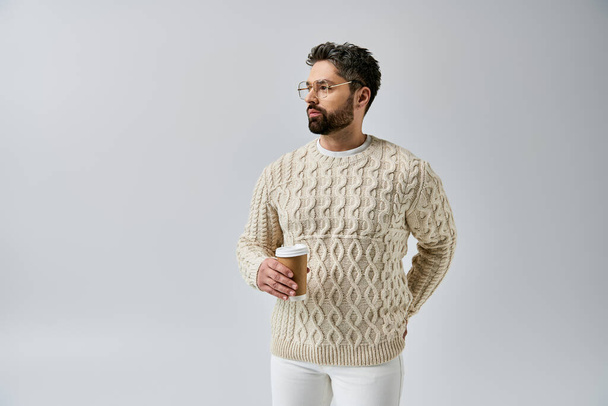 Um homem barbudo exala charme em um suéter branco contra um fundo cinza, apresentando um olhar sofisticado e polido. - Foto, Imagem