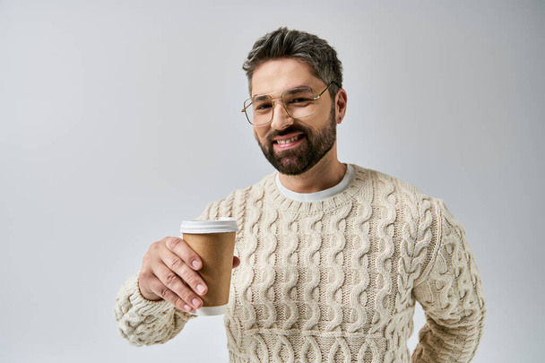 Szakállas férfi fehér pulóverben egy gőzölgő kávéscsészét tart egy hangulatos stúdióban, szürke alapon.. - Fotó, kép