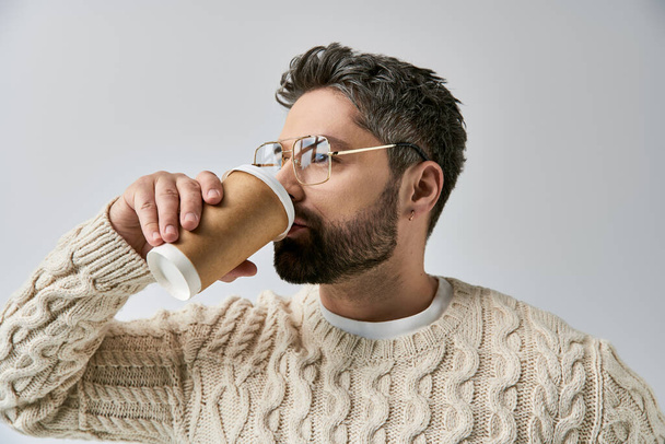 Ein bärtiger Mann im weißen Pullover, der ein Getränk aus einer Tasse genießt, während er eine Brille auf grauem Studiohintergrund trägt. - Foto, Bild