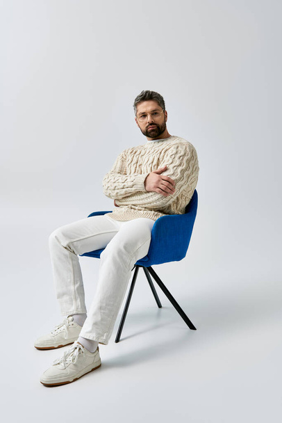Ένας γενειοφόρος άνδρας με λευκό πουλόβερ κάθεται με σταυρωμένα χέρια, αποπνέοντας γοητεία και στοχασμό σε ένα γκρίζο σκηνικό στούντιο. - Φωτογραφία, εικόνα