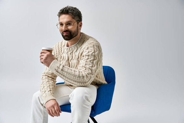 Ein Mann mit Bart sitzt in einem weißen Pullover auf einem Stuhl und hält eine Tasse Kaffee vor einem grauen Atelierhintergrund.. - Foto, Bild