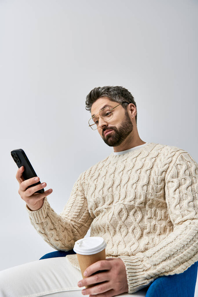 Un hombre barbudo en un suéter blanco se sienta en una silla, absorto en su teléfono celular. - Foto, Imagen