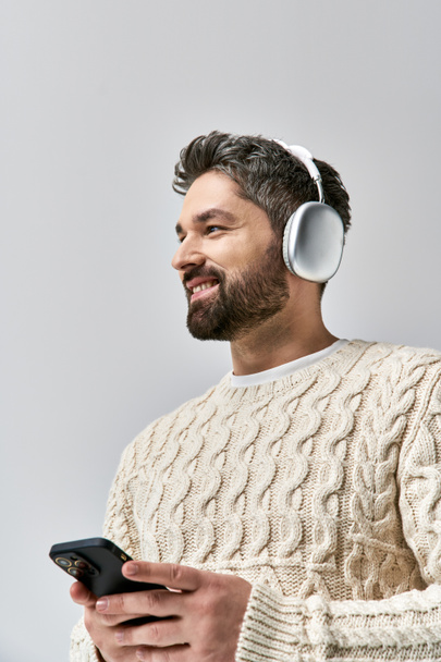 Egy szakállas férfi fehér pulóverben fejhallgatót visel, a mobilja képernyőjén egy szürke háttérrel.. - Fotó, kép