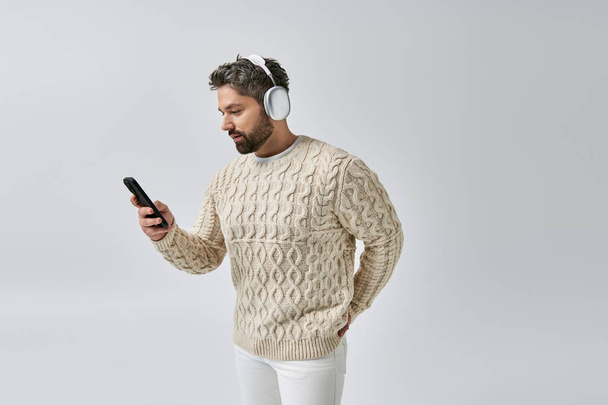 Brodaty mężczyzna w białym swetrze, noszący słuchawki, pochłonięty telefonem komórkowym na szarym tle. - Zdjęcie, obraz