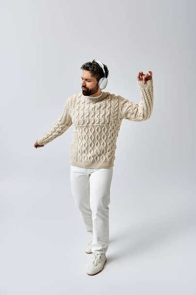 Un hombre suave y barbudo posa con un suéter blanco y pantalones contra un fondo gris del estudio. - Foto, imagen