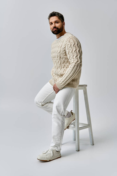 Ένας γενειοφόρος άνδρας αποπνέει γοητεία καθώς κάθεται σε ένα σκαμνί, κομψά ντυμένος με ένα λευκό πουλόβερ σε ένα γκρίζο φόντο στούντιο. - Φωτογραφία, εικόνα