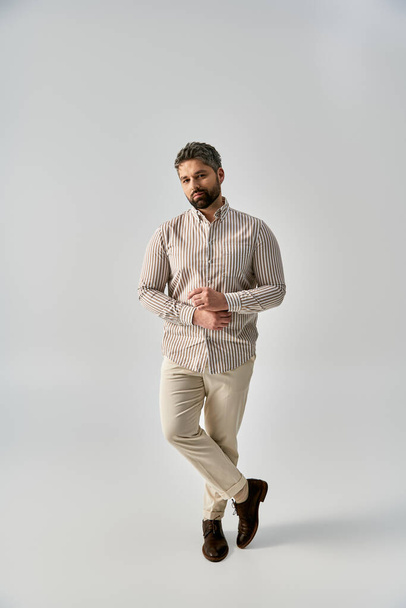 Un homme à la mode avec une barbe portant une chemise rayée et un pantalon kaki pose élégamment sur un fond gris dans un studio. - Photo, image
