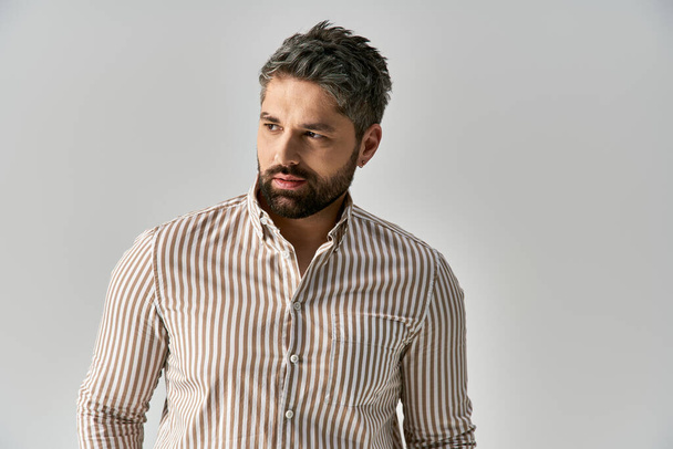Un hombre barbudo con una elegante camisa a rayas posa sobre un fondo gris de estudio. - Foto, Imagen