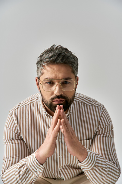 A stylish man with glasses and a beard sits gracefully, lost in thought on a grey background. - Photo, Image