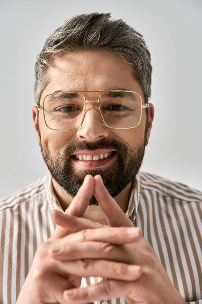 Бородатый мужчина в очках и в полосатой рубашке элегантно позирует на сером фоне в студии. - Фото, изображение