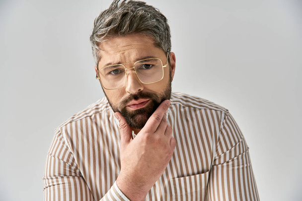 Un hombre elegante con gafas mira con confianza a la cámara con un atuendo elegante contra un fondo gris del estudio. - Foto, Imagen