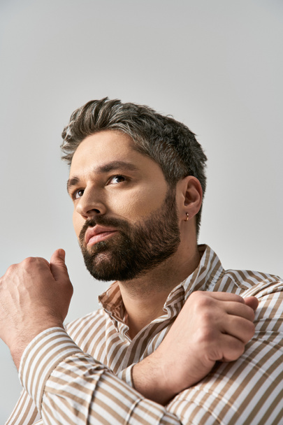 Um homem elegante com uma barba posa confiantemente em uma camisa listrada contra um fundo cinza. - Foto, Imagem