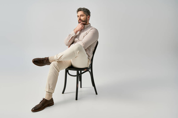 Un hombre elegante con barba y atuendo elegante se sienta en la parte superior de una silla con confianza contra un fondo gris estudio. - Foto, imagen