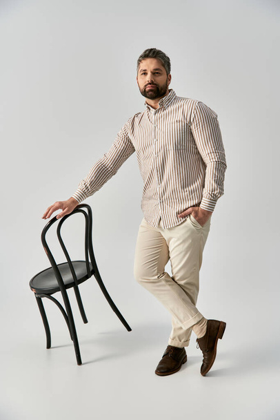 Un uomo barbuto in elegante abbigliamento colpisce una posa accanto a una sedia nera elegante su uno sfondo grigio studio. - Foto, immagini