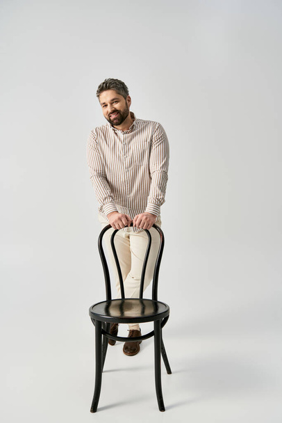 Un hombre elegante con barba está sentado en la parte superior de una silla en una pose elegante sobre un fondo gris estudio. - Foto, imagen