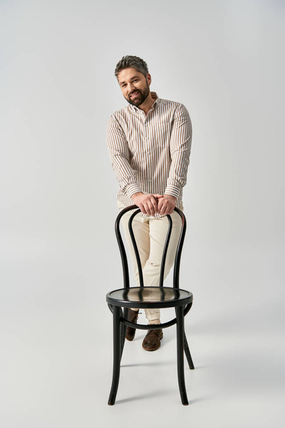 Un uomo elegante con la barba in elegante abbigliamento vicino elegante sedia nera su uno sfondo grigio neutro. - Foto, immagini