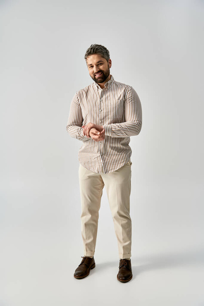 Ένας κομψός άντρας με γενειάδα ντυμένος με κομψή ενδυμασία στέκεται με αυτοπεποίθηση σε ένα γκρίζο φόντο στούντιο. - Φωτογραφία, εικόνα