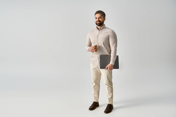 Vousatý muž v elegantním oděvu, držící notebook a šálek kávy, předvádějící rovnováhu v práci a relaxaci. - Fotografie, Obrázek
