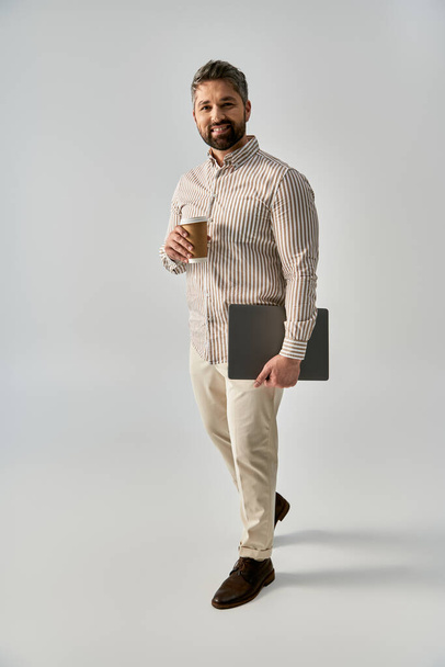 Um homem barbudo em traje elegante segurando um laptop e uma xícara de café, exalando sofisticação e relaxamento. - Foto, Imagem