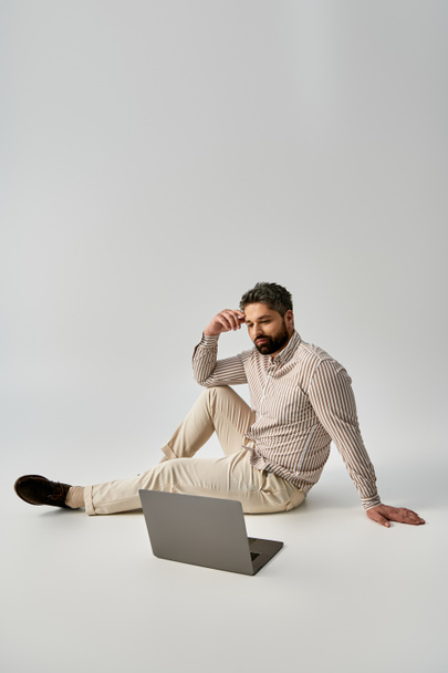 Модний чоловік з бородою сидить на землі, одягнений в елегантний одяг, оскільки він працює на своєму ноутбуці. - Фото, зображення