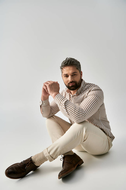 Un hombre barbudo con un atuendo elegante se sienta con las piernas cruzadas en el suelo en un entorno de estudio sobre un fondo gris, exudando aplomo. - Foto, imagen