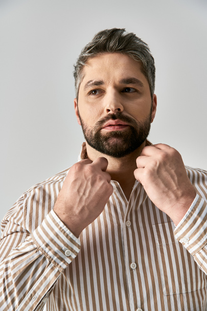 Un hombre barbudo emana confianza en una elegante camisa a rayas sobre un fondo gris de estudio. - Foto, Imagen